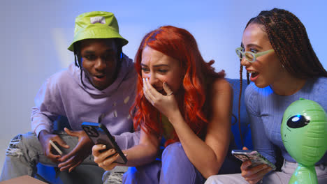 Studio-Shot-Of-Young-Gen-Z-Friends-Sitting-On-Sofa-Sharing-Social-Media-Post-On-Mobile-Phones